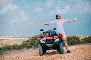 ATV 'nin yanında elinde kask olan bir adam. Beyaz elbiseli yakışıklı bir adam ve arka plandaki kumlu bir yolda çeyrek bisiklet.