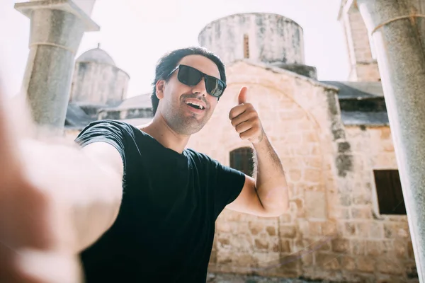 A young man takes a selfie against the background of an ancient temple on a sunny day. Handsome guy takes pictures near the ancient stone walls of the church