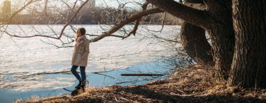 Kız baharda donmuş gölde duruyor. Erken baharda buz ile kaplı bir gölün kıyısında şehirde hafif bir yağmurluk genç kadın