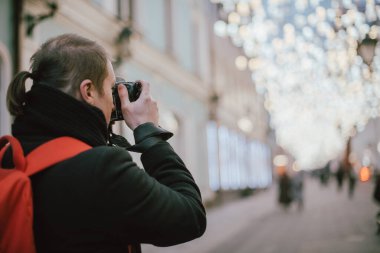 Elinde kamera olan bir adam kışın eski şehrin caddesini fotoğraflıyor. Arka planda sanatsal dekore edilmiş sokak lambalarının altında kamera ve eşarplı bir fotoğrafçı.