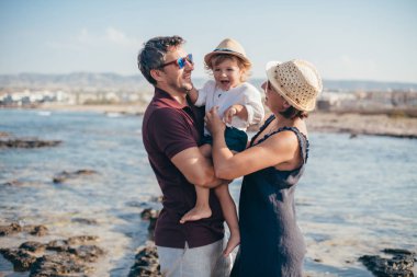 Çocuklu bir aile denizin arka planında taşların üzerinde duruyor. Şehrin arka planında bir bebek ve ufukta deniz olan genç anne ve baba.