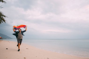 Gökkuşağı bayrağı taşıyan neşeli adam plaj boyunca koşuyor. Gökyüzü ve okyanus arka planında gökkuşağı bayrağı tutan genç adam