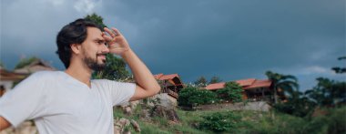 Tayland ulusal evlerinin arka planında sahilde bir erkek turistin portresi. Genç yakışıklı turist Tayland 'a seyahat ediyor.