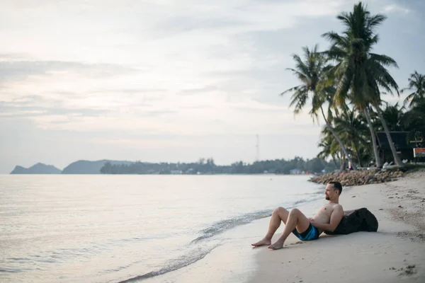 夕日のビーチで男が砂の中に横たわっている 熱帯の島で海のそばでトランクスを泳いでいる若い男 熱帯の休日 — ストック写真