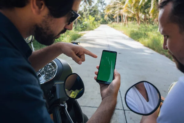 Two men on motorbikes are looking for a road on a map in a phone on a tropical island. Guys tourists are looking at the navigator on a smartphone on bikes on the road in a palm grove