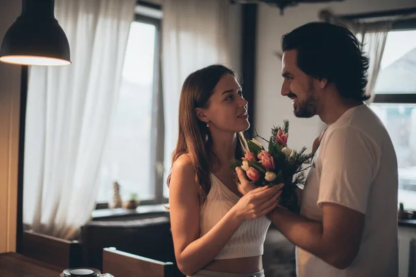 Loving couple in the evening in the living room at home. International Women\'s Day. A man gives a woman a bouquet of tulips on March 8. Romantic family evening at home.