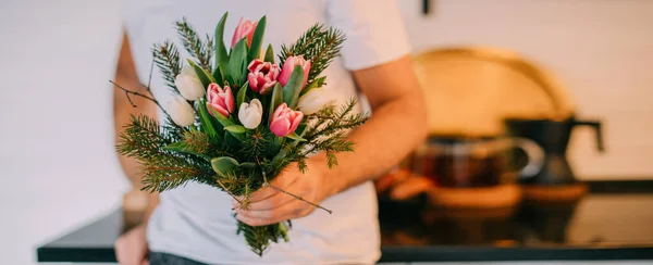 Handsome man with a bouquet in the kitchen. International Women\'s Day. A young guy holds a bouquet of tulips in his hands on March 8. Romantic family evening at home.