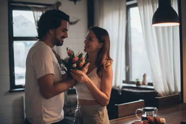 Loving couple in the evening in the living room at home. International Women's Day. A man gives a woman a bouquet of tulips on March 8. Romantic family evening at home.