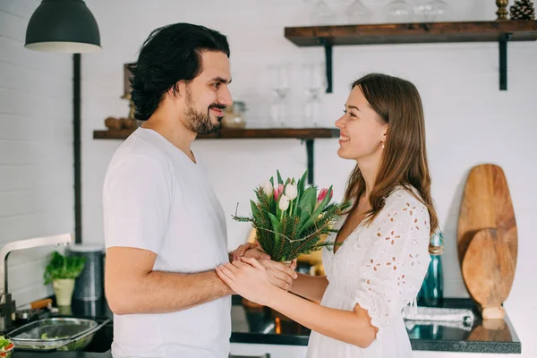 International Women's Day. Young couple in the morning in the kitchen at home. A man gives a woman a bouquet of tulips on March 8 in a beautiful stylish kitchen.