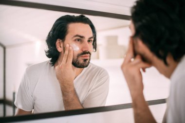 Yakışıklı adam yüz kremi sürüyor. Erkeklerin kişisel bakımı. Genç bir adam aynada kendine bakar ve yüzüne krema sürer.