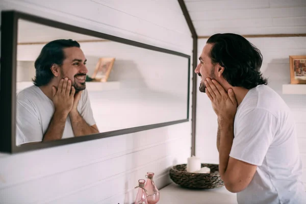 stock image A handsome man looks in the mirror. Men's personal care. A young guy examines himself in the mirror in the bedroom.