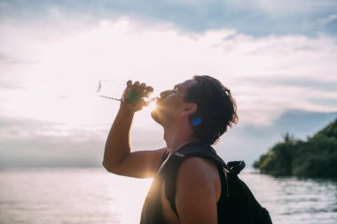 Bir adam gün batımında sahilde bir şişeden su içer. Genç bir adam deniz kenarındaki plastik bir şişeden su içiyor. Portre.