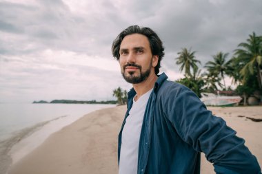 Yalnız bir adam bulutlu bir günde palmiye ağaçlarıyla kumsalda yürüyor. Genç yakışıklı adam bulutlu bir havada okyanusun kenarında kumda tek başına yürüyor..