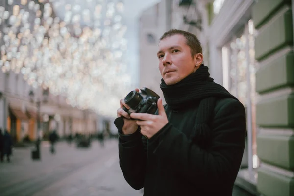 A man with a camera photographs the street of the old city in winter. Photographer with a camera in a coat and scarf on the background of artfully decorated street lamps