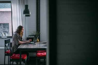 Evdeki işyerinde. Bir kadın evde dizüstü bilgisayarda çalışıyor, oturma odasındaki bir koltukta oturuyor, telefonda konuşuyor. Evden çalış..