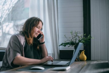 Evdeki işyerinde. Bir kadın evde dizüstü bilgisayarda çalışıyor, oturma odasındaki bir koltukta oturuyor, telefonda konuşuyor. Evden çalış..