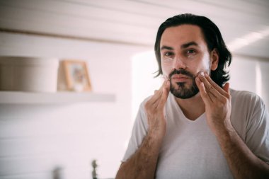 Yakışıklı adam yüz kremi sürüyor. Erkeklerin kişisel bakımı. Genç adam sabah yatak odasında yüz kremi sürüyor.