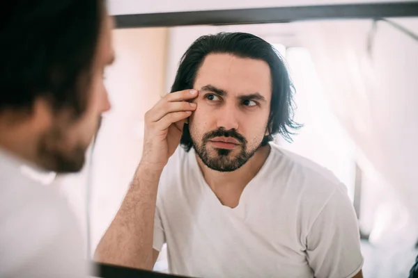 A handsome man looks in the mirror. Men's personal care. A young guy examines himself in the mirror in the bedroom.