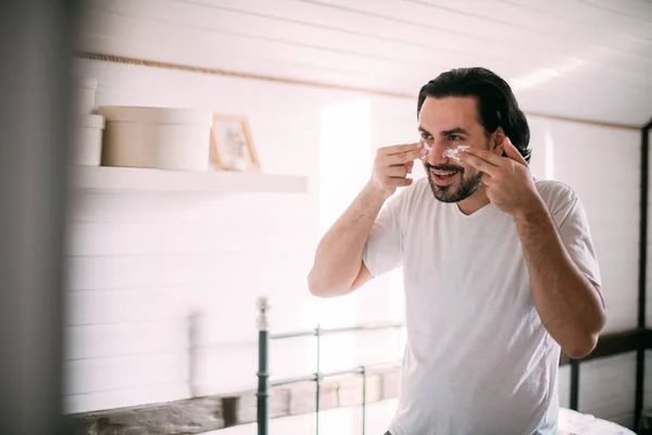 Handsome man smears face cream. Men's personal care. Young guy applies face cream in the morning in his bedroom