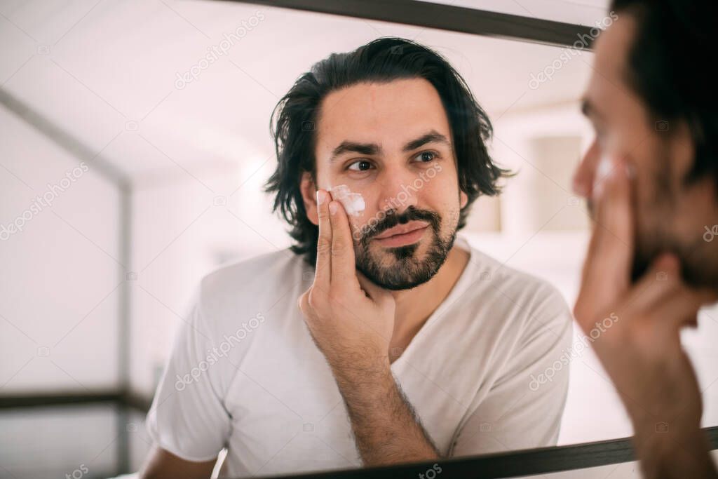 Handsome man smears face cream. Men's personal care. A young guy looks at himself in the mirror and puts cream on his face