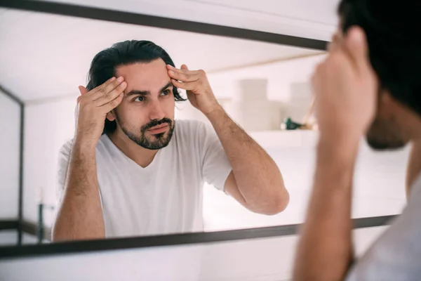 A handsome man looks in the mirror. Men's personal care. A young guy examines himself in the mirror in the bedroom.