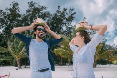 Tropik bir sahilde kokteyl içen mutlu aşıklar. Palmiye ağaçlarının, beyaz kumların ve denizin arka planında taze hindistan cevizleri olan kadın ve erkek. Tatil köyündeki balayı çifti