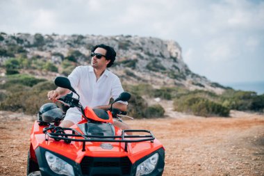 Bir adam dağlarda bir avluda oturuyor. Beyaz elbiseli yakışıklı bir adam ve kayaların arkasında kumlu bir yolda çeyrek bisiklet.