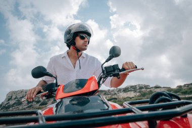Bir adam kasklı ve dağlarda gözlüklü bir avluda oturuyor. Beyaz elbiseli yakışıklı bir adam ve kayaların arkasında kumlu bir yolda çeyrek bisiklet.