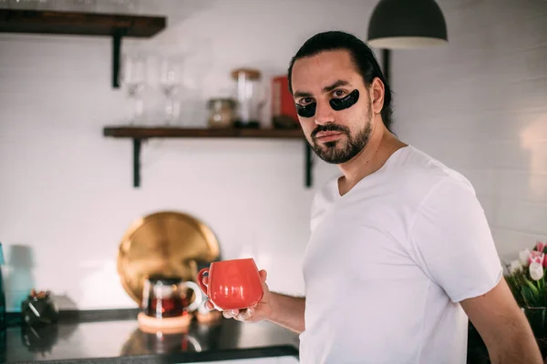 A man with patches on his face is resting. Men's personal care. Young handsome guy takes care of his face, drinks tea in the kitchen.