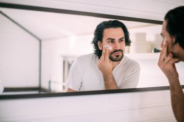 Yakışıklı adam yüz kremi sürüyor. Erkeklerin kişisel bakımı. Genç bir adam aynada kendine bakar ve yüzüne krema sürer.