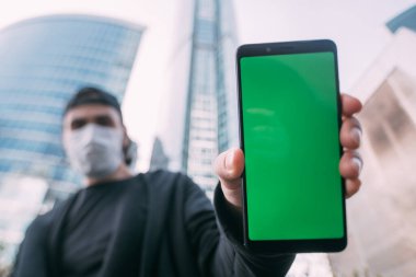 Elinde yeşil ekranı olan, maskeli bir adam. Tıbbi maskeli genç bir adam ön planda gökdelenlerin arka planında bir telefon tutuyor.