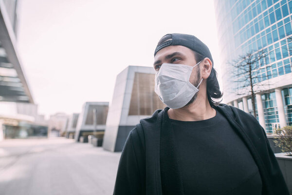 A man in a medical mask on an industrial background of skyscrapers. Lonely young guy in a mask in an empty metropolis. Man and huge city in quarantine.