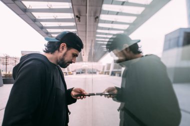 Elinde telefonu ve yansıması olan bir adam. Hayatın çevrimiçi dönüşümü. Genç bir adam, bir megalopolis 'in camından endüstriyel bir arka plana yansıyor..