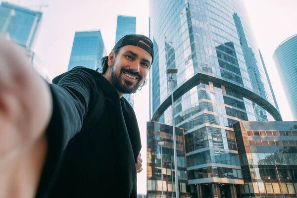 A man on an industrial background of glass skyscrapers takes a selfie. A lonely young guy in a metropolis. Man and a huge city.