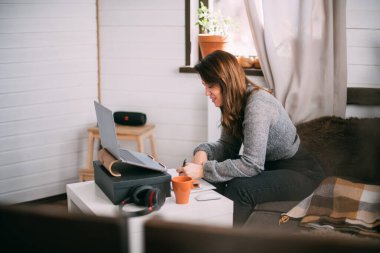 Bir kadın oturma odasında dizüstü bilgisayarla oturuyor, ekrana bakıyor, notlar alıyor, internetten yabancı bir dil öğreniyor, online eğitim kursları okuyor, internetten eğitim alıyor, bir müşteriye danışmanlık yapıyor.