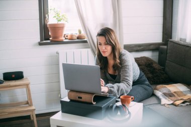 Bir kadın oturma odasında dizüstü bilgisayarla oturuyor, ekrana bakıyor, notlar alıyor, internetten yabancı bir dil öğreniyor, online eğitim kursları okuyor, internetten eğitim alıyor, bir müşteriye danışmanlık yapıyor.