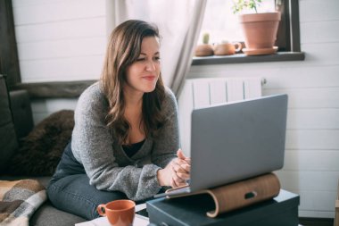 Bir kadın oturma odasında dizüstü bilgisayarla oturuyor, ekrana bakıyor, notlar alıyor, internetten yabancı bir dil öğreniyor, online eğitim kursları okuyor, internetten eğitim alıyor, bir müşteriye danışmanlık yapıyor.