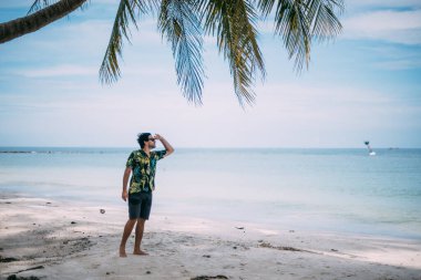 Hawaii gömlekli güzel bir adam tropik bir plajda yürüyor. Adam tropikal bir adadaki palmiye ağaçlarının arka planında okyanusa yaklaşır.