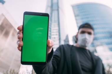 Elinde yeşil ekranı olan, maskeli bir adam. Tıbbi maskeli genç bir adam ön planda gökdelenlerin arka planında bir telefon tutuyor.