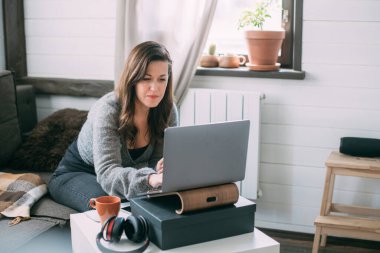 Bir kadın oturma odasında dizüstü bilgisayarla oturuyor, ekrana bakıyor, notlar alıyor, internetten yabancı bir dil öğreniyor, online eğitim kursları okuyor, internetten eğitim alıyor, bir müşteriye danışmanlık yapıyor.