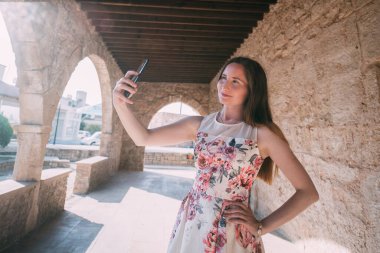 Genç bir kadın güneşli bir günde antik bir tapınağın arka planında selfie çekiyor. Kilisenin antik taş duvarlarının yanında bir kızın fotoğrafı çekiliyor..