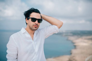 Denize bakan kayalıklarda bir adam. Beyaz elbiseli yakışıklı adam kayalık sahildeki okyanus manzarasının tadını çıkarıyor. # Denizin ötesindeki genç adam #