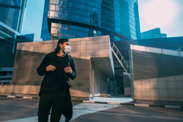 A man in a medical mask on an industrial background of skyscrapers. Lonely young guy in a mask in an empty metropolis. Man and huge city in quarantine.