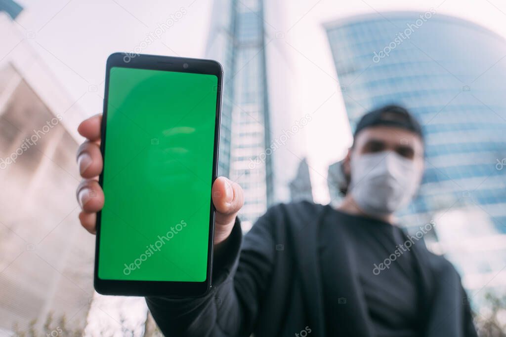 A man in a mask with a phone with a green screen in his hands. A young guy in a medical mask holds a phone in the foreground against the background of skyscrapers