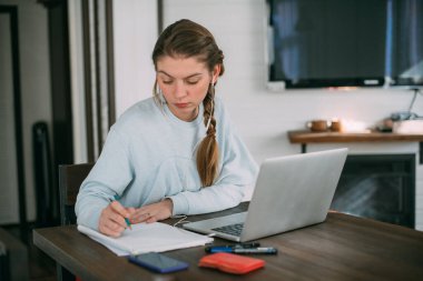 Bir kadın oturma odasında dizüstü bilgisayarla oturuyor, ekrana bakıyor, notlar alıyor, internetten yabancı bir dil öğreniyor, online eğitim kursları okuyor, internetten eğitim alıyor, bir müşteriye danışmanlık yapıyor.