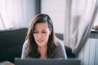 Bir kadın oturma odasında dizüstü bilgisayarla oturuyor, ekrana bakıyor, notlar alıyor, internetten yabancı bir dil öğreniyor, online eğitim kursları okuyor, internetten eğitim alıyor, bir müşteriye danışmanlık yapıyor.