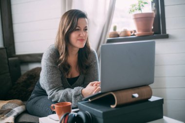 Bir kadın oturma odasında dizüstü bilgisayarla oturuyor, ekrana bakıyor, notlar alıyor, internetten yabancı bir dil öğreniyor, online eğitim kursları okuyor, internetten eğitim alıyor, bir müşteriye danışmanlık yapıyor.