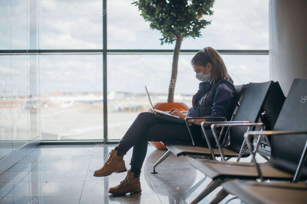 A woman is sitting at the airport with a laptop in a medical mask. A young girl is waiting for departure at the gate, working online. Social responsibility, virus protection.