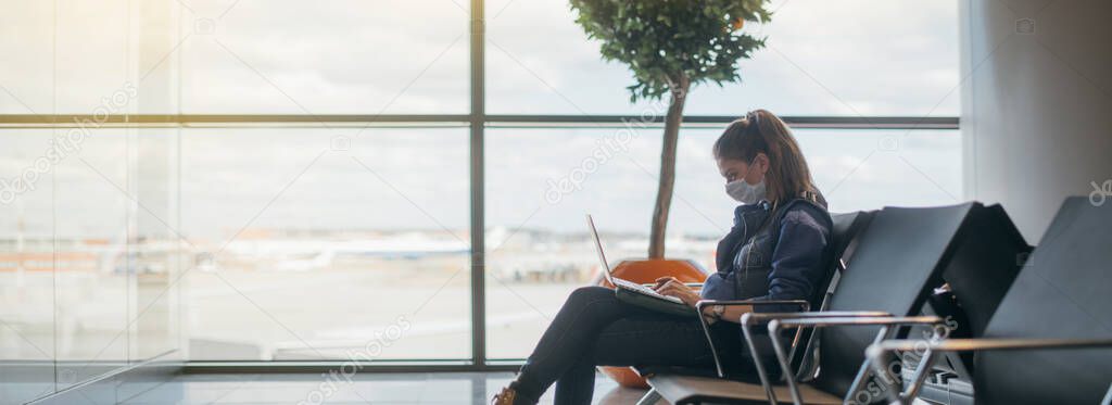 A woman is sitting at the airport with a laptop in a medical mask. A young girl is waiting for departure at the gate, working online. Social responsibility, virus protection.