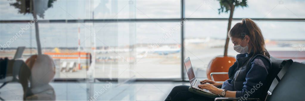 A woman is sitting at the airport with a laptop in a medical mask. A young girl is waiting for departure at the gate, working online. Social responsibility, virus protection.